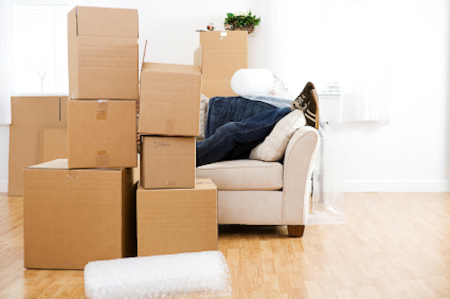 Umzugskartons stehen vor einer Couch und bedecken eine Person bis zur Hälfte, die darauf liegt und ihre erste eigene Wohnung gemietet hat.
