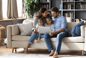 eine junge Familie sitzt auf einer weißen Couch vor einem Laptop und lächeln.