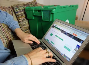 geöffneter Laptop auf dem gearbeitet wird. Dahinter steht eine grüne Umzugsbox und grün-rote Kissen